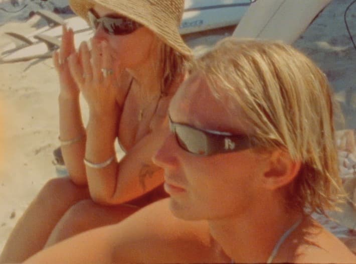 Two people sit close together on the sand in sunglasses, one in a straw hat, boards stacked behind them.