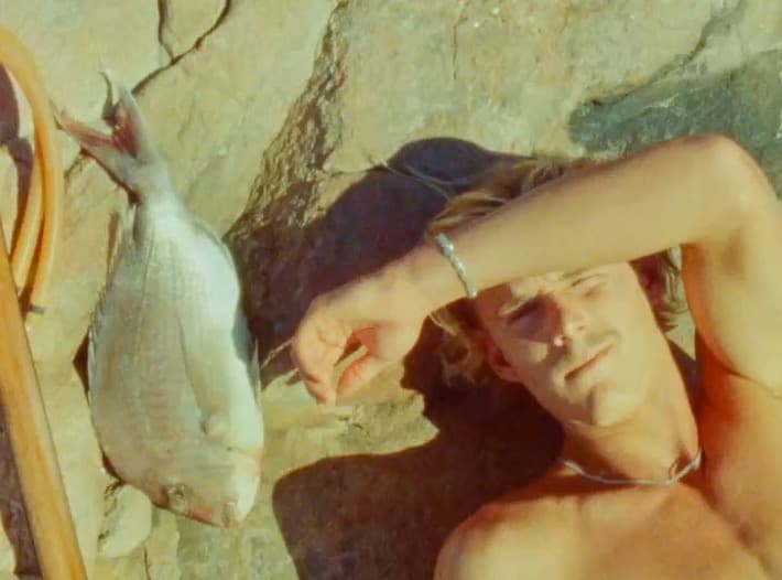 A spearfisher lies back on warm rock with an arm across his brow, his catch and speargun laid out beside him.