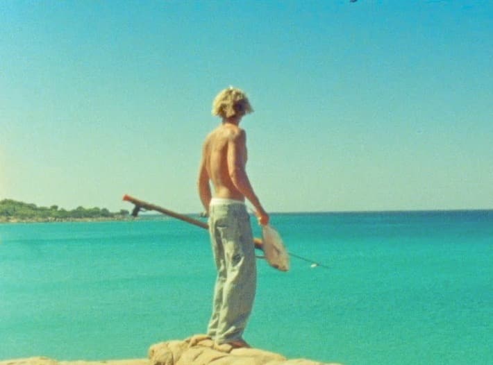 A spearfisher stands on a rock ledge with his back to camera, speargun under one arm, looking out across flat turquoise water.