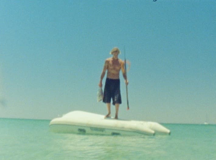 A spearfisher in a wide-brimmed hat stands on a half-submerged dinghy in pale green shallows, spear in one hand and a fish in the other.