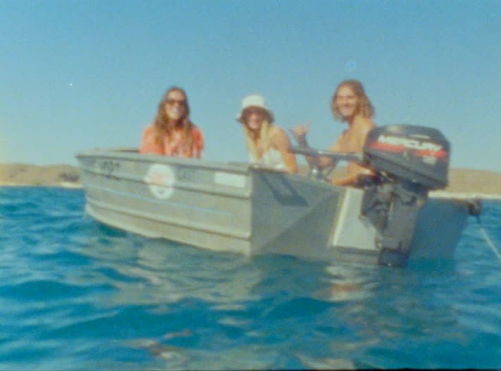 Three friends sit in a small aluminium boat on flat turquoise water, one throwing a shaka from the outboard, low red dunes behind them.